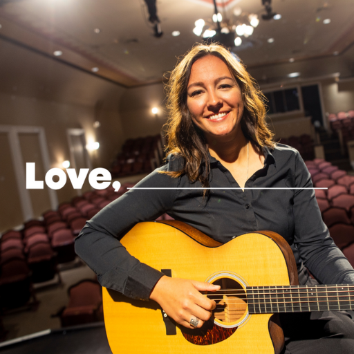 A smiling woman with blonde hair is wearing a black long-sleeved shirt and holding a guitar. Behind her is rows of burgundy theatre seats. 